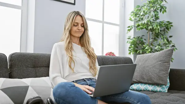 stock image A blonde woman in casual wear using a laptop on a couch at home, showcasing everyday technology use in a living room.