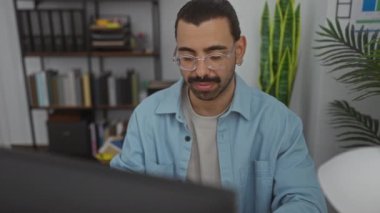 Ofiste çalışan genç bir adam, gülümseyen ve gözlük takan, bilgisayarın önünde, arka planda bitki ve raflarla, rahat bir atmosferi olan kapalı bir çalışma alanını gösteriyor.