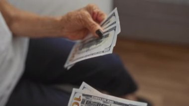 Yaşlı bir kadın evde otururken birden fazla 100 dolarlık banknot tutuyor. Finansal güvenlik ve kıdemli yaşam tarzını ön plana çıkarıyor..