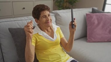 Oturma odasındaki kanepede oturan, elinde uzaktan kumandayla gülümseyen, kısa saçlı yaşlı İspanyol kadın..