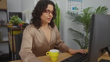 Genç bir İspanyol kadın kollarını uzatıyor ve kapalı ofis ortamında yorgun görünürken gözlüğünü çıkarıyor..