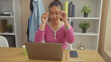 Ofiste dizüstü bilgisayarda çalışan gözlüklü, bitki ve raf geçmişi olan İspanyol bir kadın..