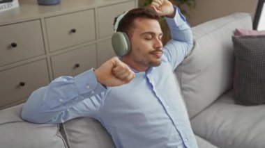 Rahat genç adam, rahat bir oturma odasında kulaklıkla müzik dinliyor..