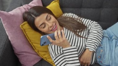 Tatlı bir odada renkli yastıkların üzerinde uzanmış akıllı telefon kullanan İspanyol kadın gülümsüyor.