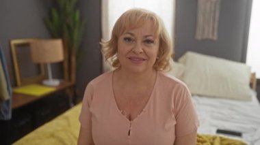 Orta yaşlı sarışın kadın gülümseyerek yatakta oturuyor, dişlerini gösteriyor ve mutlu, pozitif ve kendinden emin görünüyor.