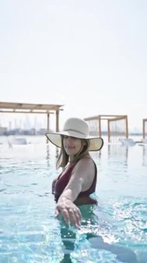 Smiling woman in swimsuit and sunhat lounging at luxury poolside with clear blue water.