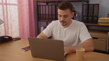 Genç bir İspanyol, ofis ortamında dizüstü bilgisayarı, kahve fincanı ve telefonuyla çalışıyor. Organizasyon dosyalarıyla çevrili..