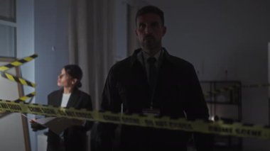 Dedektifler karanlık bir suç mahallini ellerinde belgeler tutan bir kadın ve dikkat bandının arkasında duran bir adamla birlikte inceliyorlar..