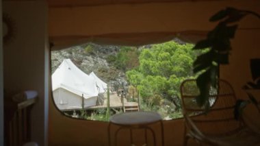 Bir pencere çerçevesinden görünen Afrika doğasında büyüleyici bir çadır, yemyeşil ağaçlar, davetkar bir ahşap güverte, ve kırsal dekor, sakin bir kamp macerasını çağrıştıran.