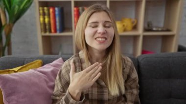 Kanepede oturan öfkeli sarışın genç kadın, elleriyle havayı kesiyor boğazına bıçak tehdidi, saldırganlık ve şiddet belirtisi olarak.