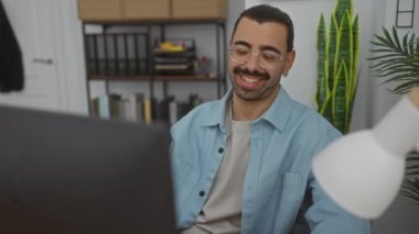 Bıyıklı ve gözlüklü bir genç açık mavi bir gömlek giyip arka planda bitkiler ve kitap raflarıyla bilgisayar ekranına bakarken gülümsüyor.