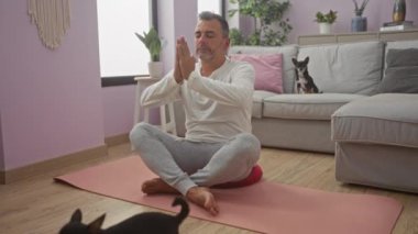 İspanyol bir adam, rahat bir oturma odasında pembe bir paspasın üzerinde yoga yapıyor. Kanepede iki tane chihuahua var. Etrafı ev bitkileri ve dekorla çevrili..
