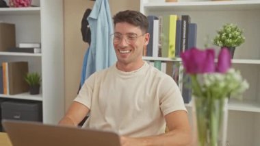Modern bir ev ofisinde dizüstü bilgisayarıyla meşgul olan neşeli bir genç adam, yaşam tarzını ve uzaktan çalışmayı yansıtıyor..