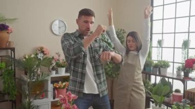 Bir erkek ve bir kadın neşeyle dans ediyorlar, güneşli, bitki dolu bir çiçekçide, kapalı bir eğlence aktivitesi sergiliyorlar..