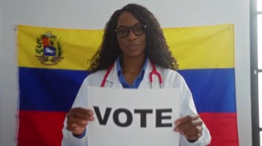 Kıvırcık saçlı ve gözlüklü Afro-Amerikan bir kadın, Venezuela bayrağının önünde elinde bir oy pusulası tutarak seçim kampanyasında oy vermeye teşvik ediyor..