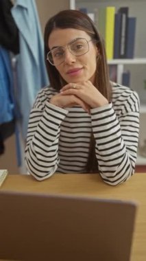 Ofiste dizüstü bilgisayarı ve kitaplığı olan çizgili tişörtlü, gülümseyen İspanyol kadın..