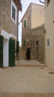Valldemossa, Mallorca Limanı 'nda geleneksel taş kaplı bir sokakta yürüyen, siyah bir elbise giyen ve güneşli bir günde kamış çantası taşıyan bir kadın..