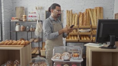 Fırında bıyıklı genç İspanyol adam akıllı telefon kullanıyor. Etrafı ekmek ve hamur işleriyle çevrili.