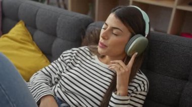 Rahatlamış bir İspanyol kadın, rahat bir oturma odasında kulaklıklarla müzik dinlemekten keyif alır. Dinginlik ve gündelik bir tarz yayar..