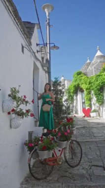 İtalya 'nın Alberobello şehrinde yeşil elbiseli genç İspanyol kadın parlak çiçekler ve açık mavi gökyüzünün altında resimli taş trulli evlerle süslenmiş bisikletiyle dikiliyor..