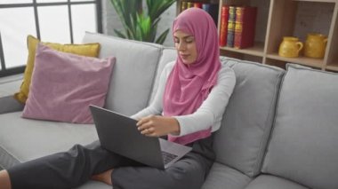 Tesettürlü bir kadın, rahat ve rahat bir oturma odasında, gri bir koltukta oturan bir dizüstü bilgisayarla dikkatle çalışır..