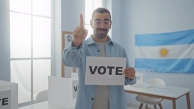 Bıyıklı genç adam Arjantin Seçim Koleji 'nde kapalı mekanlarda bayrak ve oy kullanma kabinleriyle oy kullanıyor.