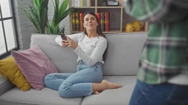 İspanyol kadın ve erkek, rahat bir oturma odasında kanepede kahve ve akıllı telefondan keyif alıyorlar.