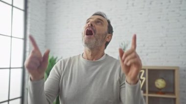 Neşeli orta yaşlı adam, şaşırmış, şaşkınlık ve mutluluğu işaret ediyor! Evde süveter giyer, kendine güveni tamdır, evde neşe dolu bakar..
