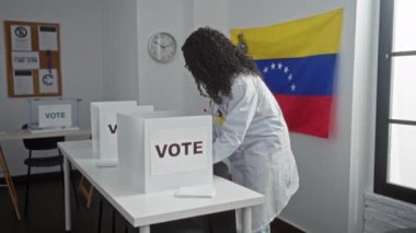 Sağlık ceketli genç bir Afro-Amerikalı kadın, arka planda Venezuela bayrağı olan bir üniversite odasında oy verirken görülüyor..