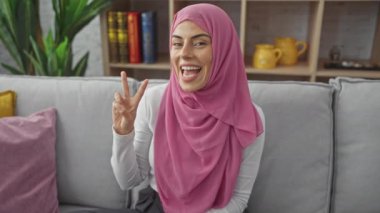 Tesettürlü neşeli, genç İspanyol kadın evde büyük kazanıyor, gülümsüyor, göz kırpıyor ve rahat koltuğundan zafer işareti gösteriyor.