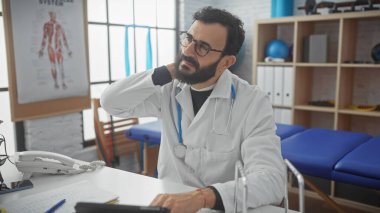Beyaz laboratuvar önlüklü, sakallı olgun bir adam, kapalı bir klinikte, boynuna dokunan düşünceli bir doktor gibi davranıyor..