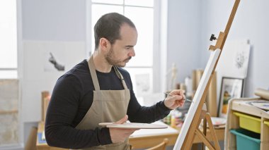 Parlak sanat stüdyosundaki tuvale odaklanmış sakallı adam önlük giyiyor.