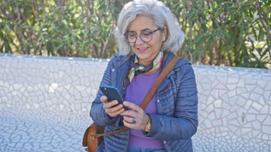 Güneşli parkta akıllı telefon kullanan, gülümseyen ve teknolojiden zevk alan olgun gri saçlı bir kadın..