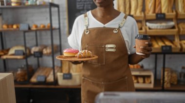 Genç Afro-Amerikalı kadın bir fırının içinde kahve ve hamur işleri tutuyor, arka planda ahşap raflar bulunan çeşitli ekmek ve fırınlanmış ürünleri sergiliyor, kahverengi önlük takıyor.