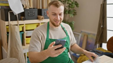 Önlüklü sakallı adam atölyedeki akıllı telefonu neşeyle kullanıyor.