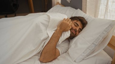 Latin asıllı genç bir adam, yatak odasında beyaz çarşaflarla uzanıyor. Sakallı çekici bir yetişkinin evinde huzur içinde uyumasının keyfini çıkarıyor..
