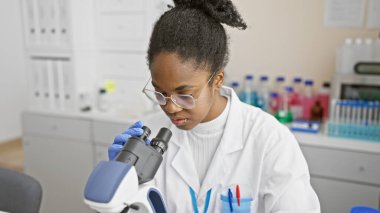 Afrikalı Amerikalı kadın bilim adamı bir laboratuarda mikroskopla örnekleri analiz ediyor.