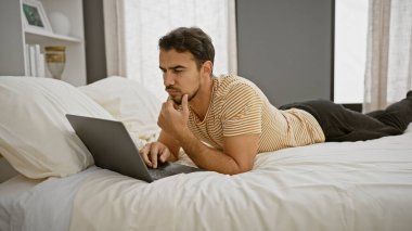 Yatakta sakallı, modern bir yatak odasında dizüstü bilgisayar kullanan Latin kökenli genç bir adam..