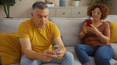 Orta yaşlı bir adam ve kadın oturma odasında akıllı telefonlar kullanarak bir koltukta oturuyor ve modern ve rahat bir ev atmosferini sergiliyorlar..