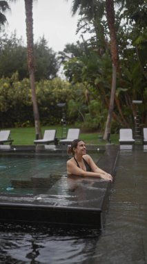 Dalgın genç bir kadın, bereketli bir bali tatil köyünün açık havuzunda sükunetin tadını çıkarıyor. Rahatlık ve lüksün vücut bulmuş hali..