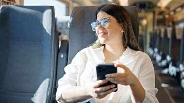 Genç, güzel, İspanyol bir kadın gülümsüyor. Akıllı telefon kullanıyor. Trende oturuyor.