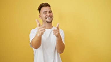 Sakallı, gülümseyen ve sarı arka planda kamerayı işaret eden genç İspanyol adam.