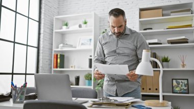 Olgun sakallı bir adam modern ofis ortamında belgeleri inceliyor, profesyonellik ve deneyim yayıyor..