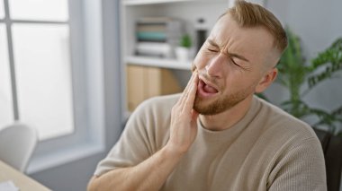 Yetişkin bir beyaz adam ofiste acı içinde kıvranıyor, parlak, modern bir iç mekanın arka planına karşı çenesini tutuyor..