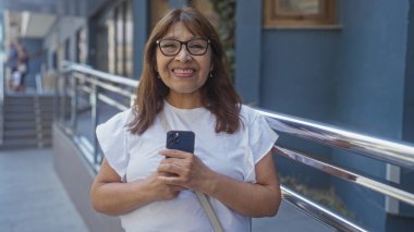 Güzel, orta yaşlı, İspanyol bir kadın. Güneşli bir günde arka planda kentsel binalar olan, gülümseyen ve elinde akıllı bir telefonla şehir sokaklarında dikiliyor..
