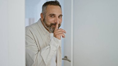 A mature hispanic man with a beard and grey hair gestures silence at the entrance of a home. clipart