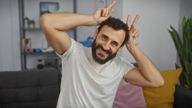 Sakallı bir adam oturma odasında gülümserken tavşan kulağı hareketi yapıyor..