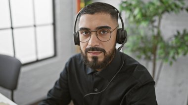 Profesyonel ofis ortamında kulaklık ve gözlük takan sakallı genç bir adam odaklanmış ve dikkatli görünüyor..