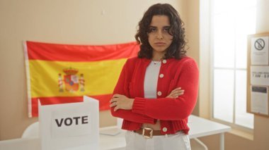 Çapraz kollu genç İspanyol kadın, İspanyol bayrağı ve oy verme kabiniyle bir üniversite odasında duruyor..
