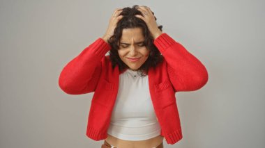 Genç İspanyol bir kadın hayal kırıklığına uğramış görünüyor, elleri başının üstünde, beyaz bir arka planda izole edilmiş kırmızı hırka ve beyaz gömlek giyiyor..